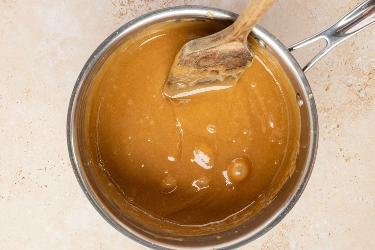 Golden brown penuche mixture bubbling in a stainless steel saucepan with a wooden spoon resting inside.