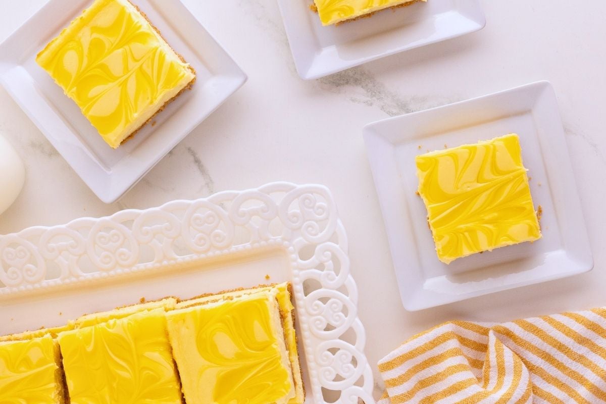 Bright yellow swirled lemon cheesecake bars on white square plates and a decorative tray with striped napkin.