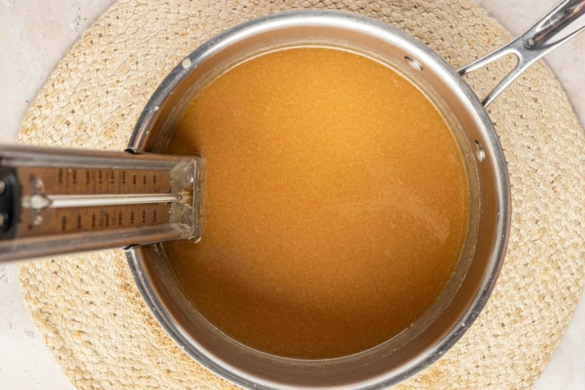 Amber sugar syrup cooling undisturbed in a stainless saucepan with a candy thermometer clipped to the side.
