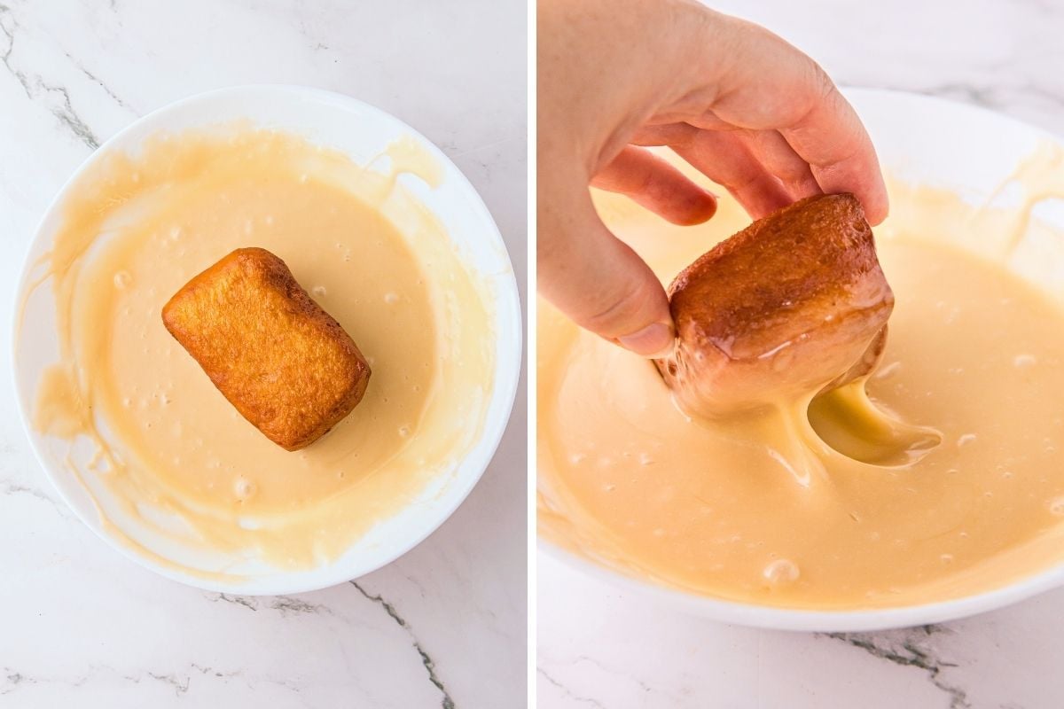 Golden fried donut being dipped into a white bowl of glossy maple glaze on marble.