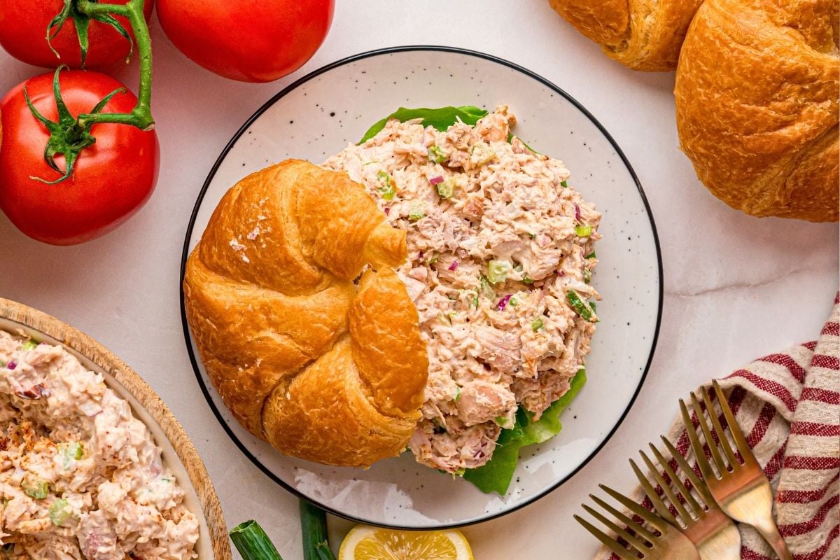 Creamy smoked chicken salad served on a flaky croissant with lettuce, tomatoes, and forks nearby.