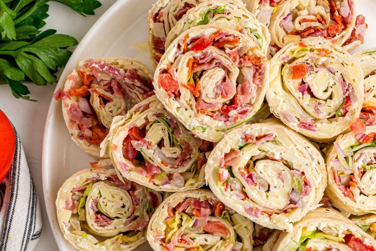 Italian ham pinwheels stacked on a white platter with fresh parsley and a striped napkin nearby.