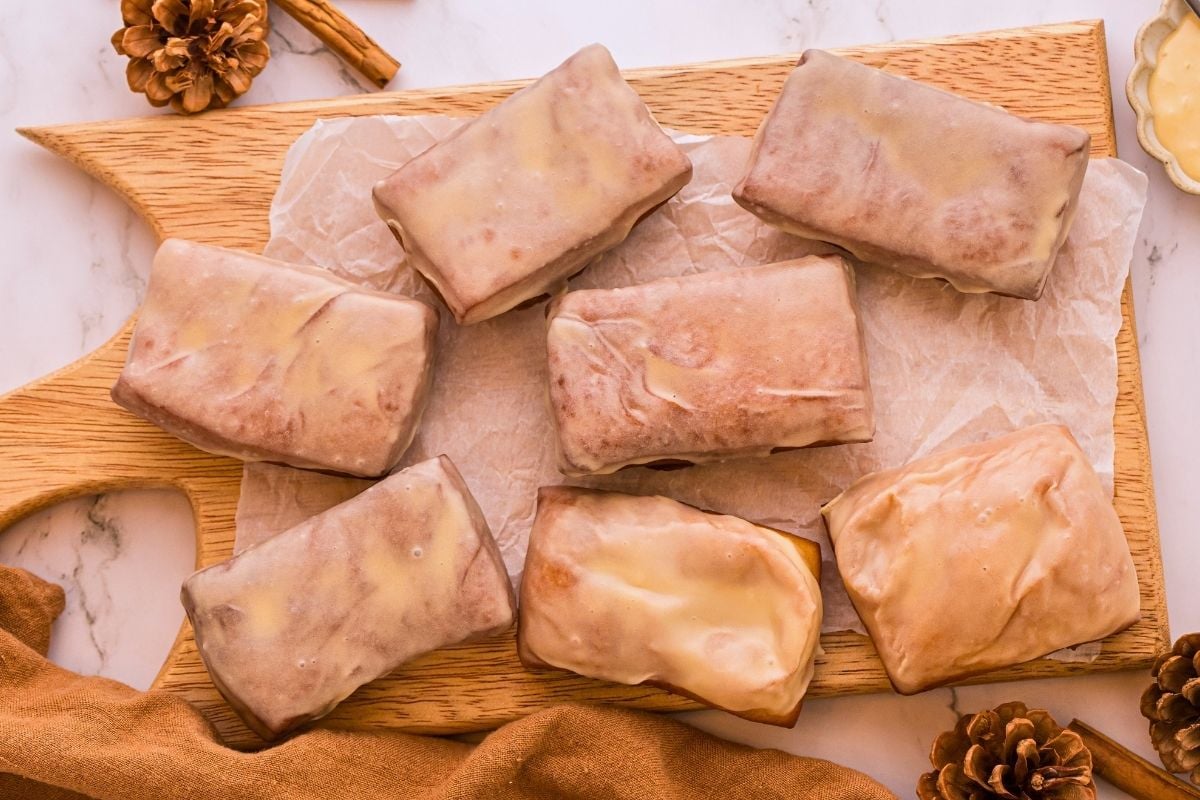 Seven maple-glazed bar donuts arranged on parchment paper atop a wooden board with pinecones and cinnamon sticks nearby.
