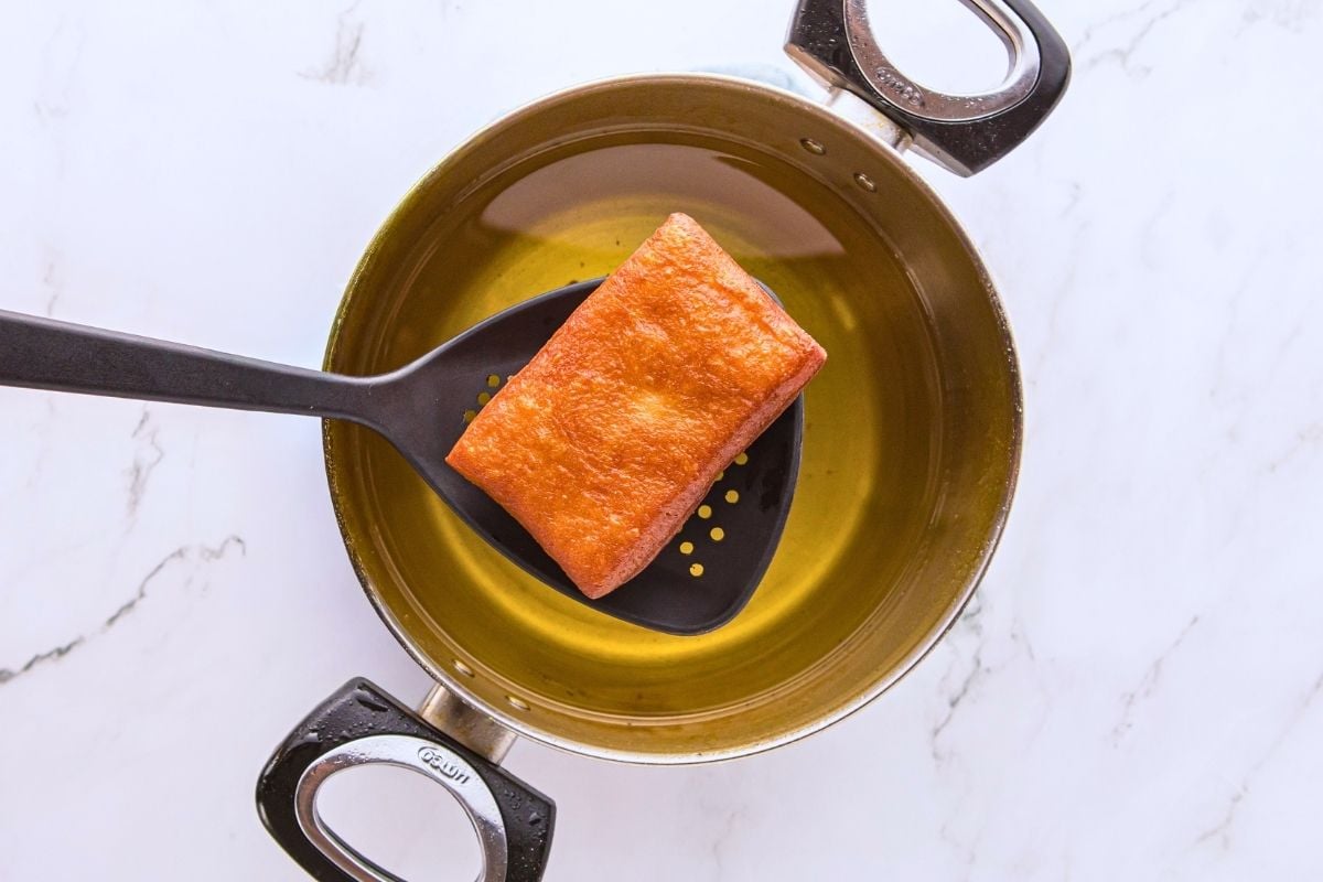 Golden fried donut bar lifted from a pot of hot oil with a black slotted spoon.