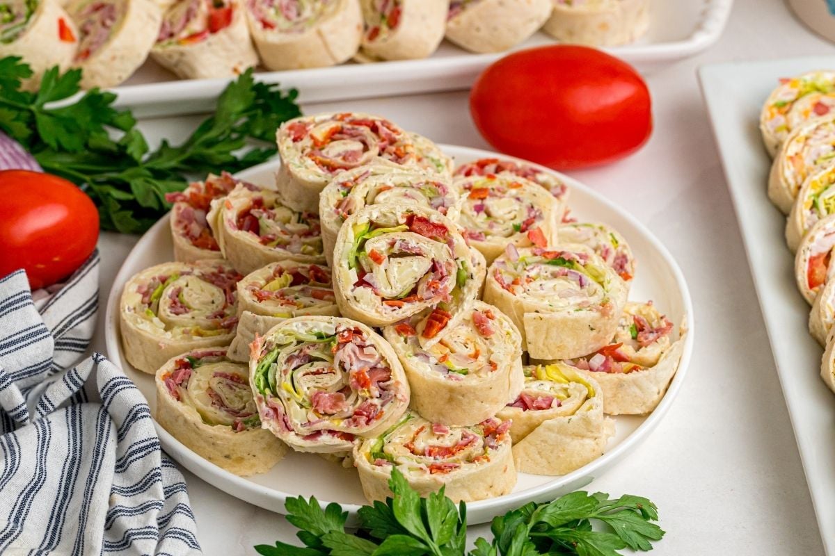Italian pinwheels stacked on a white plate with fresh parsley, ripe tomatoes, and a striped napkin nearby.