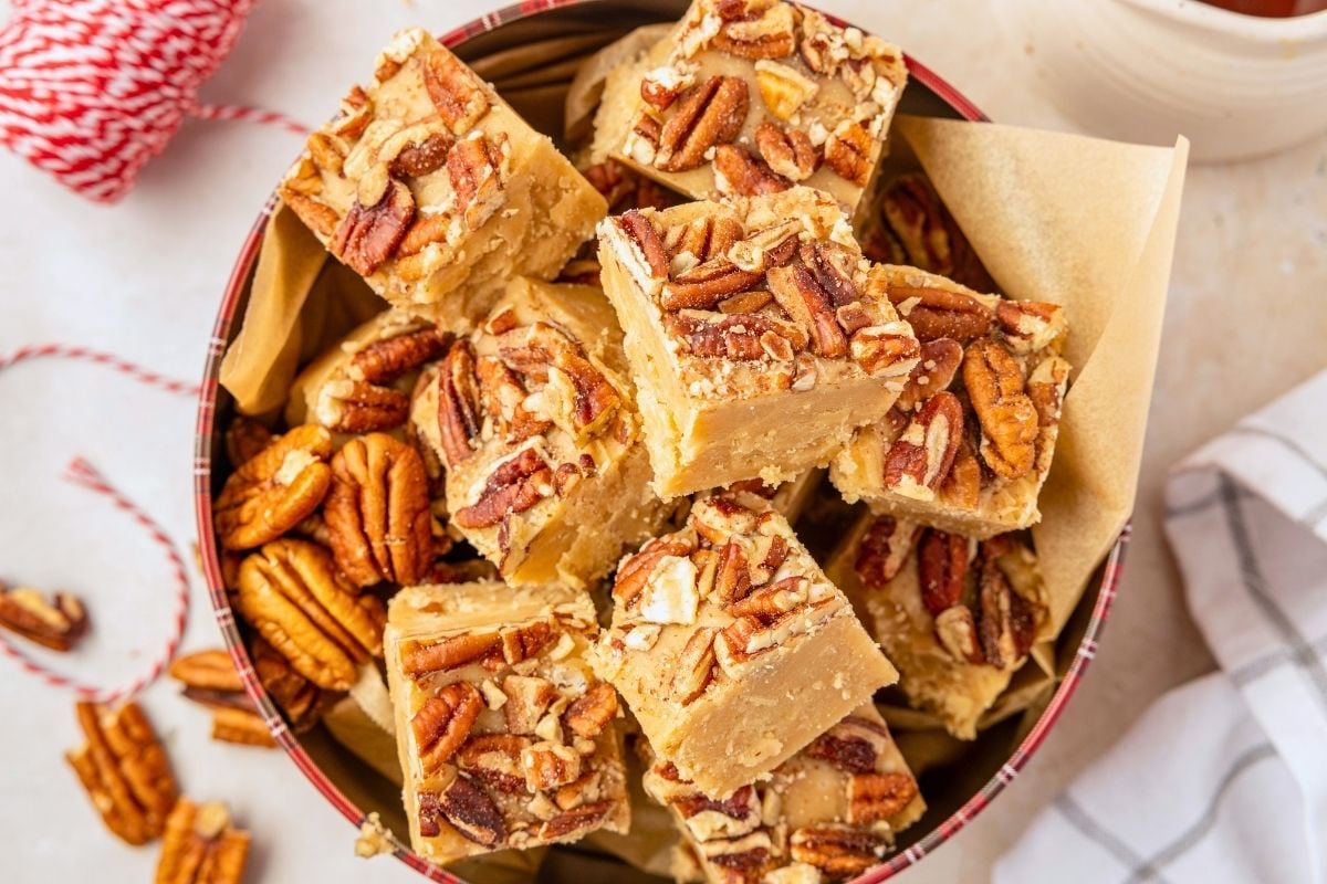 Squares of penuche fudge topped with chopped pecans piled in a parchment-lined tin beside baker's twine.