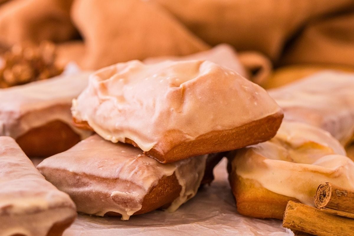 Stack of glazed maple bar donuts on parchment paper with cinnamon sticks and warm tan cloth backdrop.