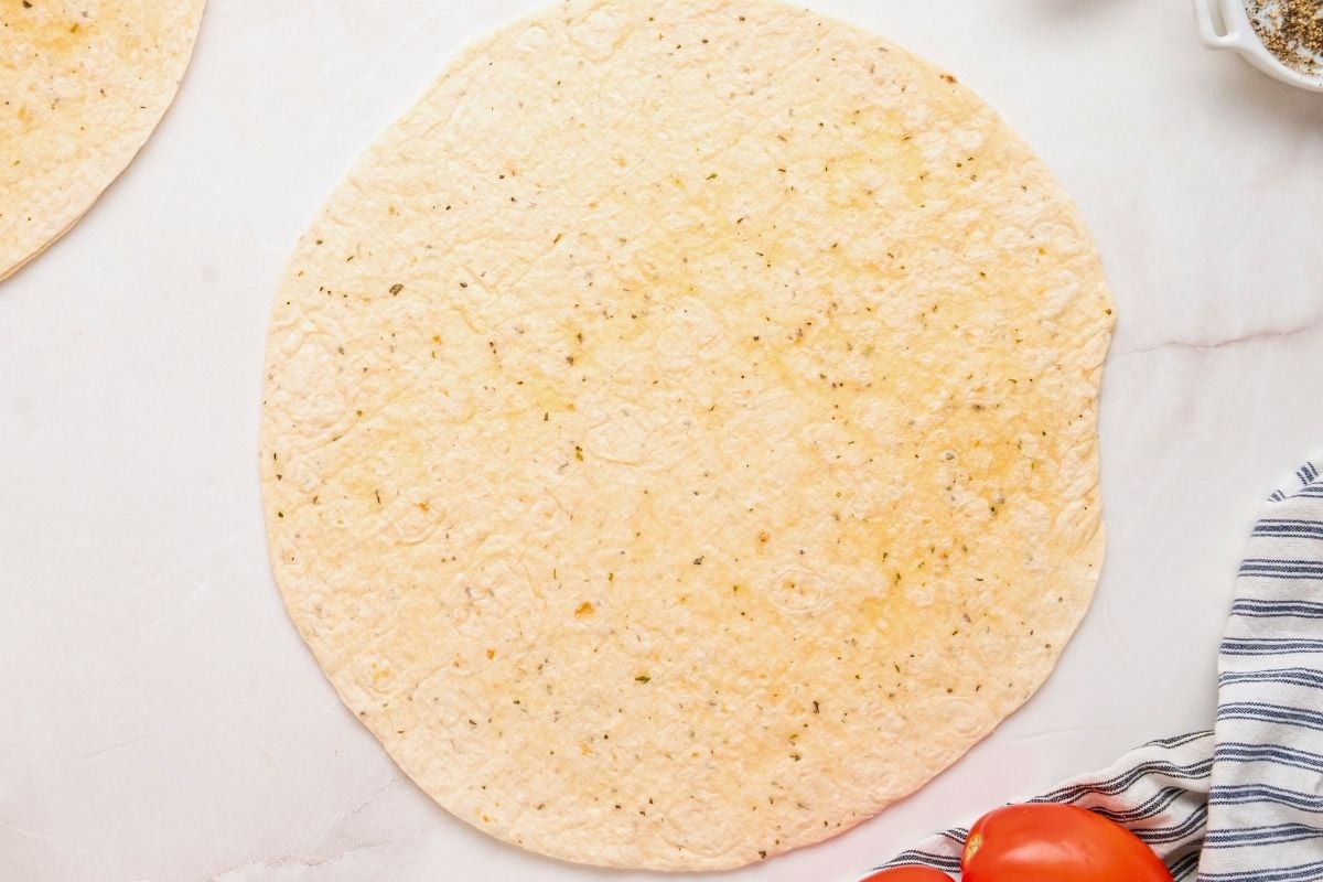 Herb-flecked tortilla laid flat on a white counter beside a striped towel and tomato.
