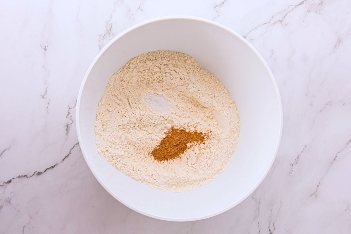 Flour, baking powder, and cinnamon combined in a large white mixing bowl on marble.