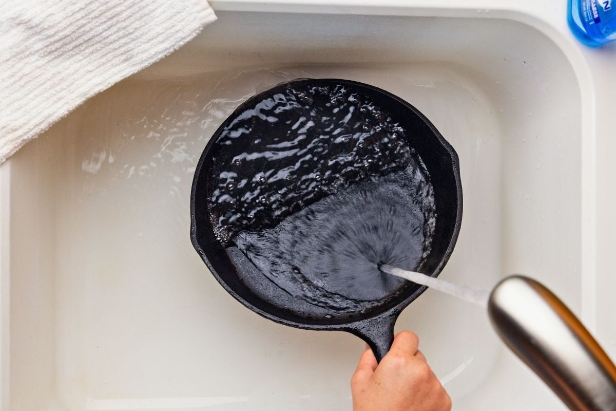 Hand rinsing a black cast iron skillet under running faucet water in a white sink.