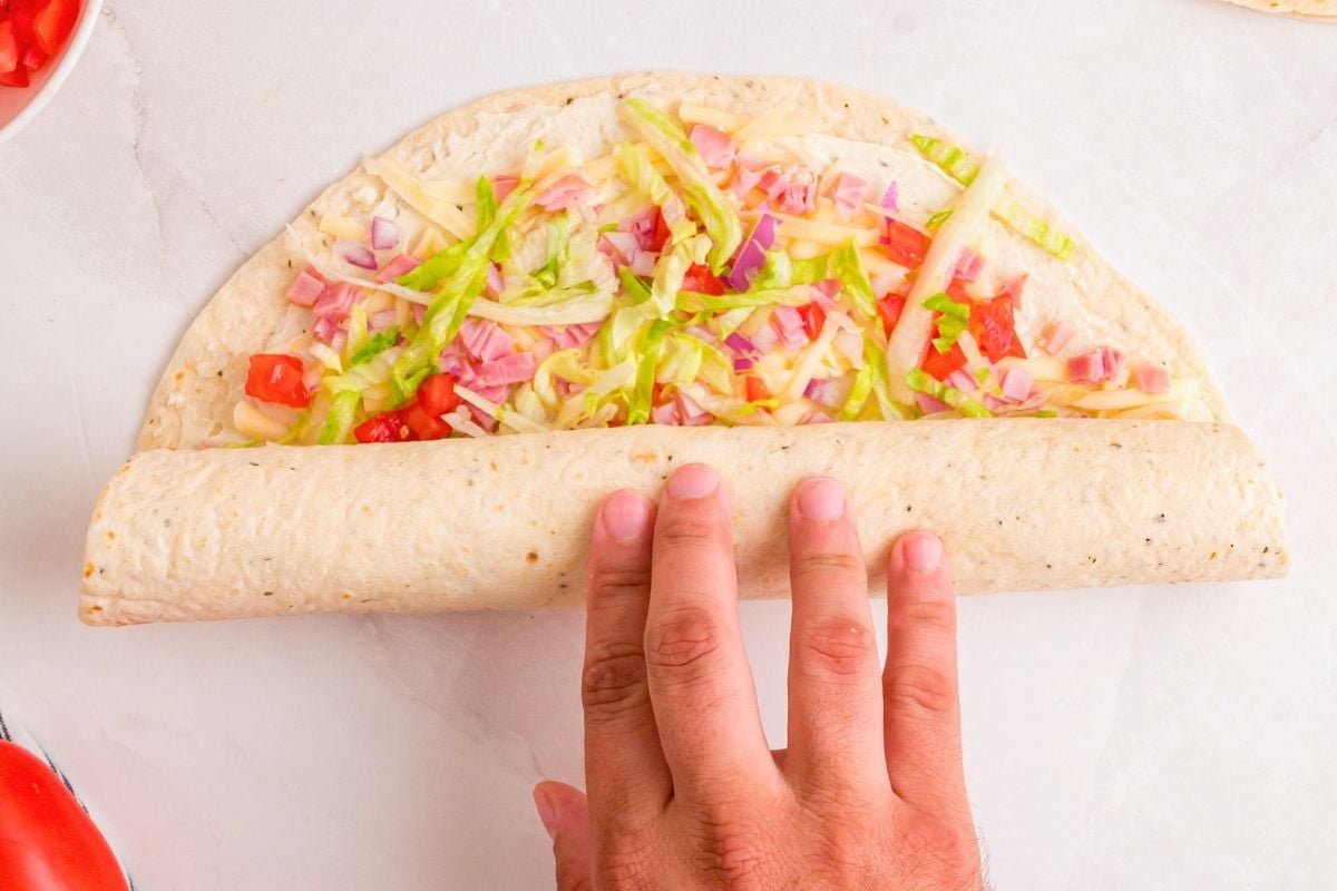 Hand rolling up a tortilla filled with shredded lettuce, ham, cheese, and diced tomatoes.