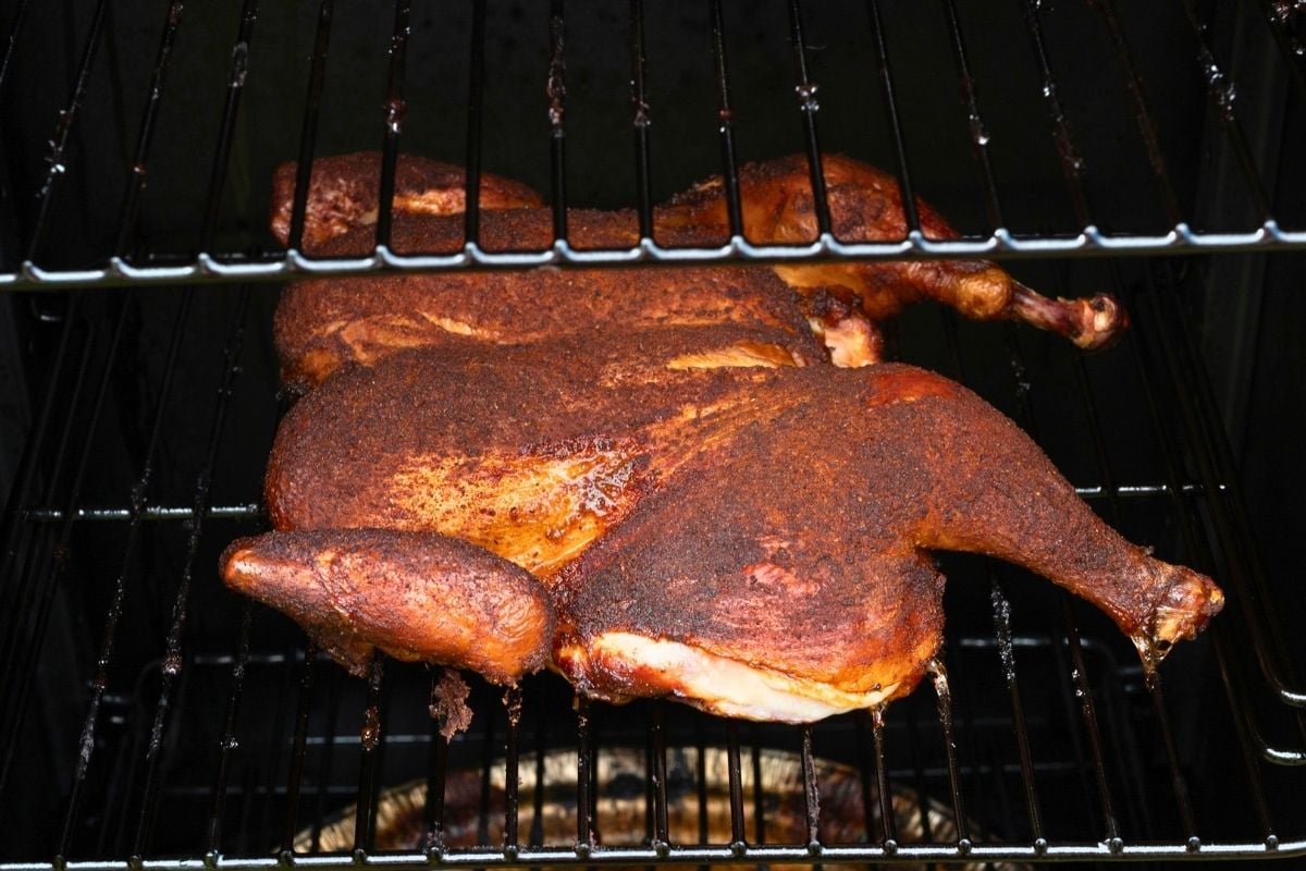 Two spatchcocked chickens with deep mahogany spice rub cooking on the metal grates of a smoker.