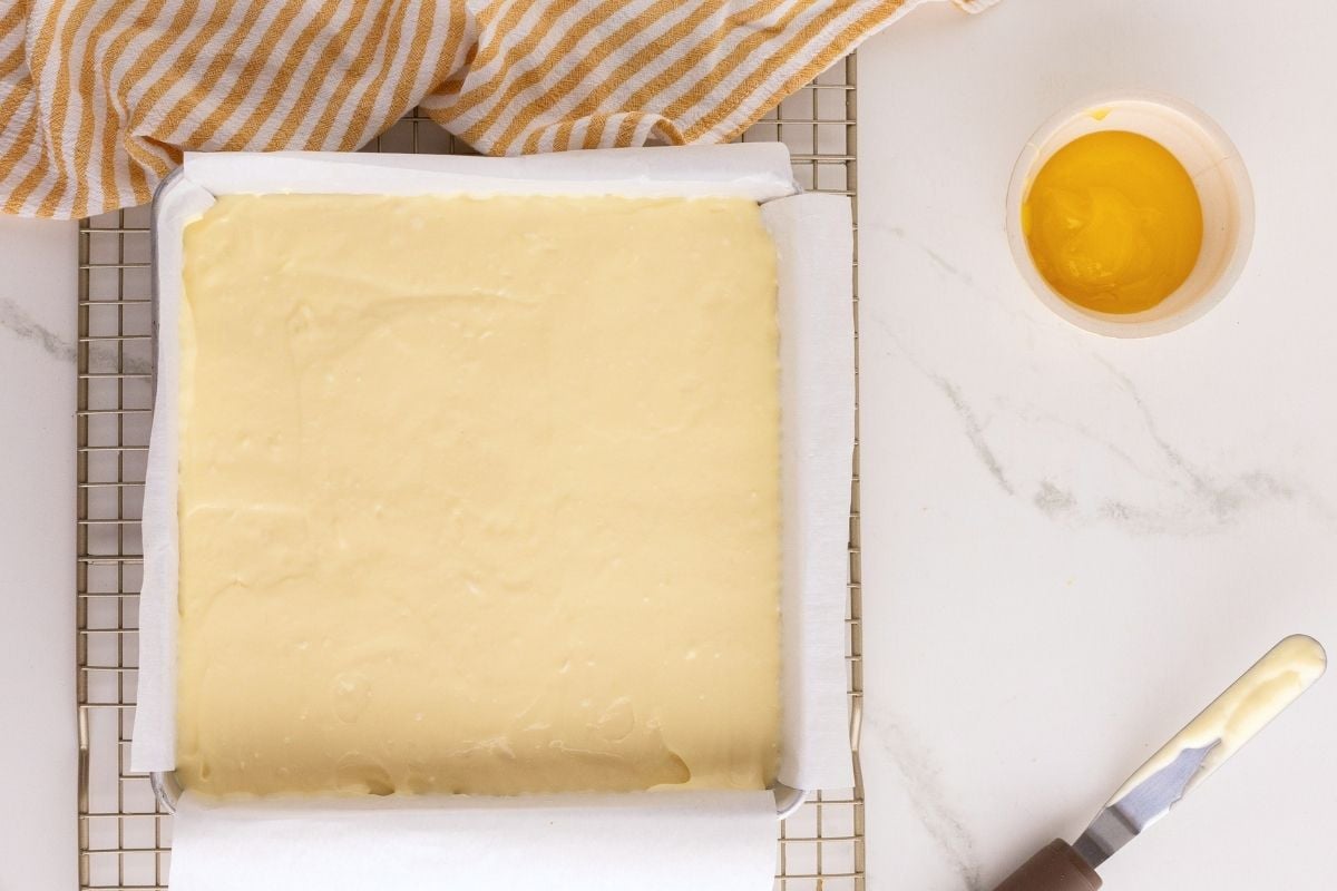 Creamy cheesecake batter poured into a parchment-lined square pan beside a small bowl of lemon curd.