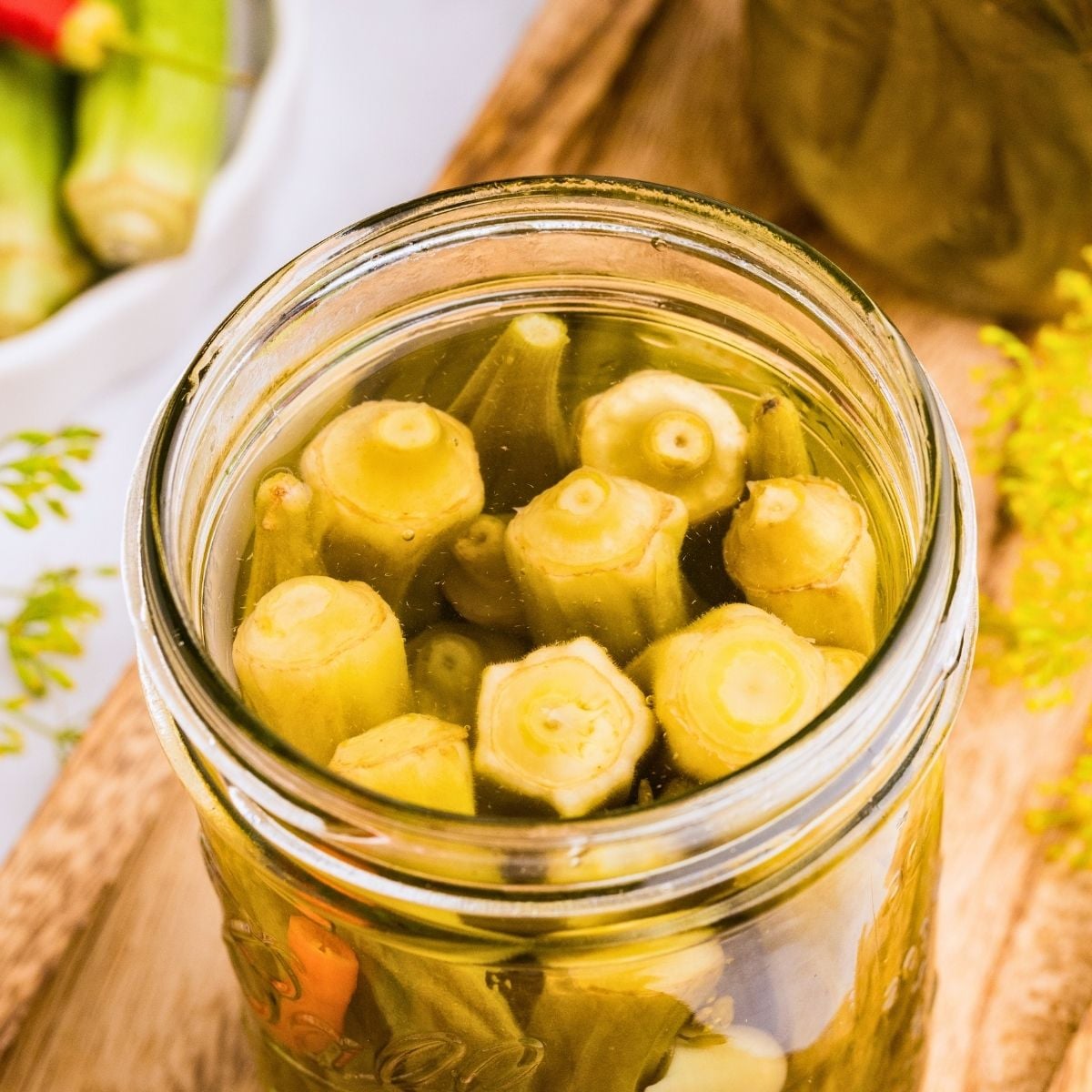 Open glass mason jar packed with pickled okra pods in brine on a wooden cutting board.