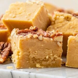 Squares of creamy caramel-colored penuche fudge topped with chopped pecans on a marble surface.