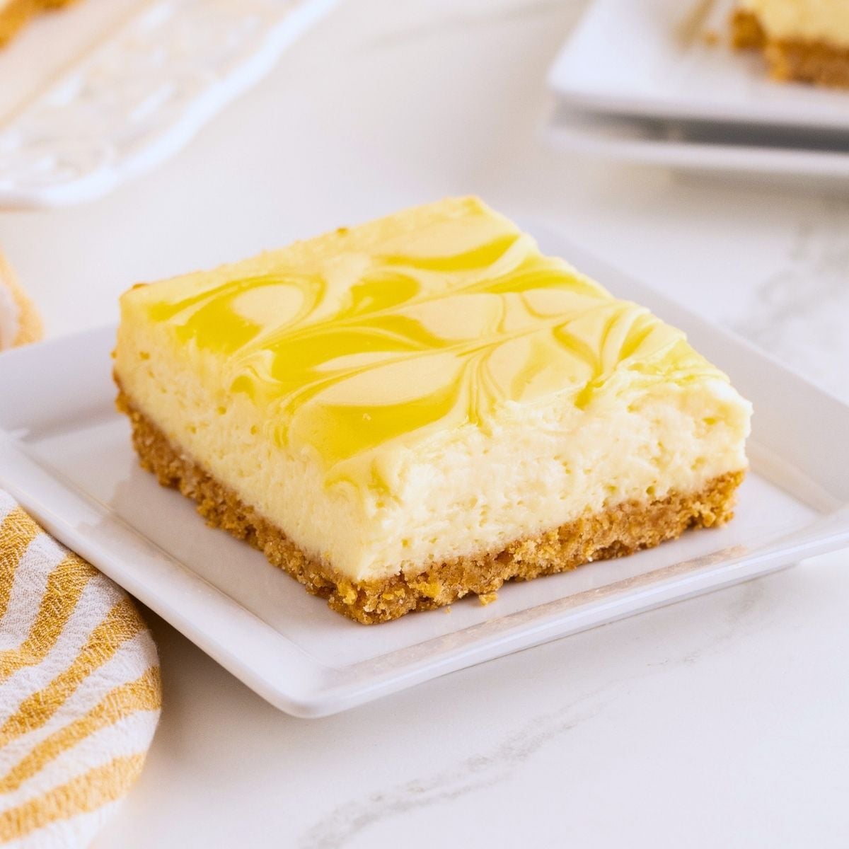 Square lemon cheesecake bar with yellow swirled topping and graham cracker crust on a white plate.