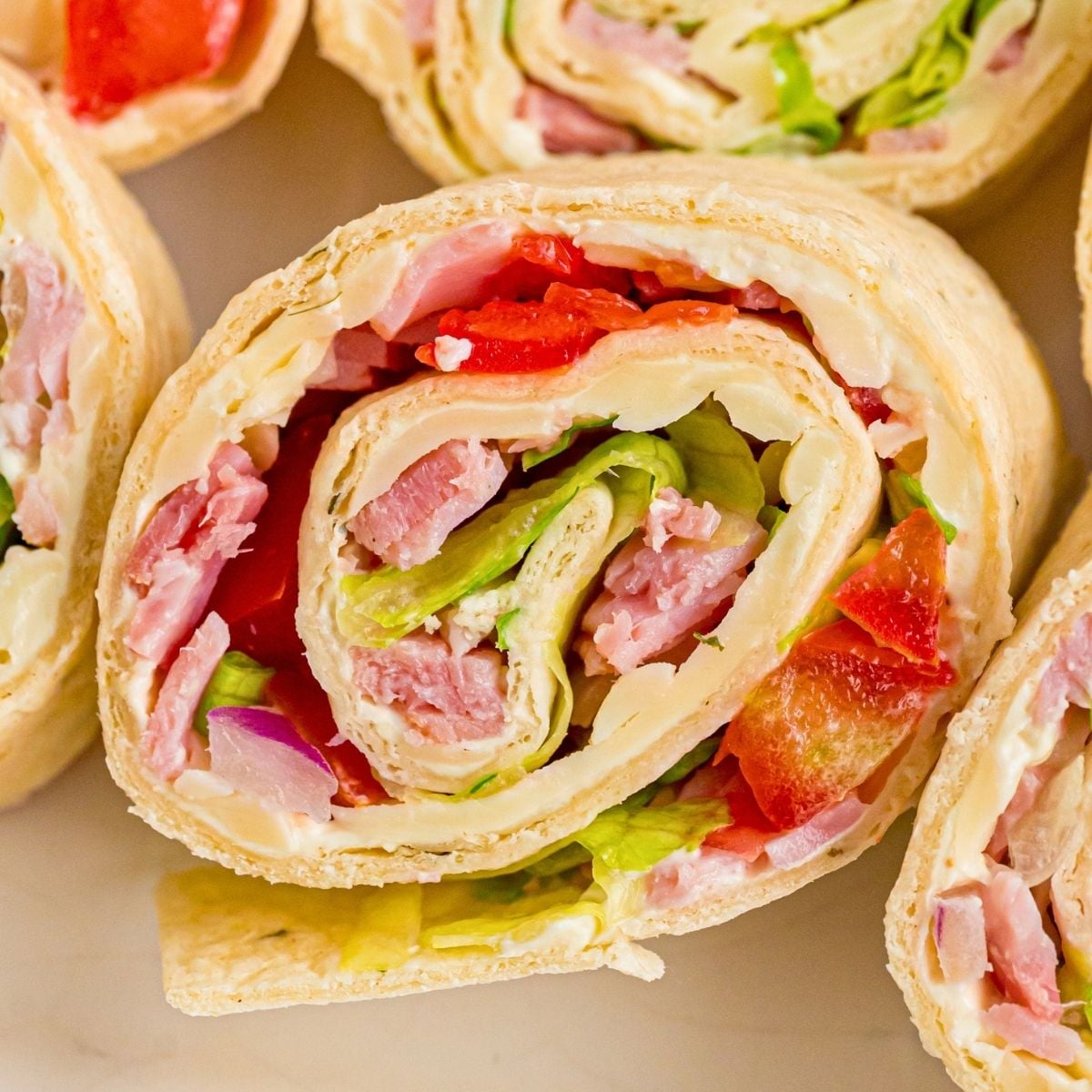 Close-up of tortilla pinwheels filled with ham, lettuce, red peppers, onion, and cream cheese on a light surface.