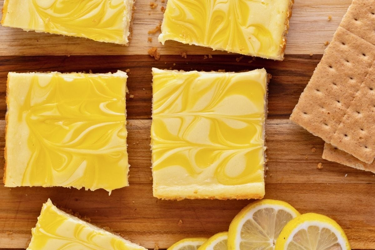 Squares of lemon cheesecake bars with yellow swirled tops on a wooden board beside graham crackers and lemon slices.