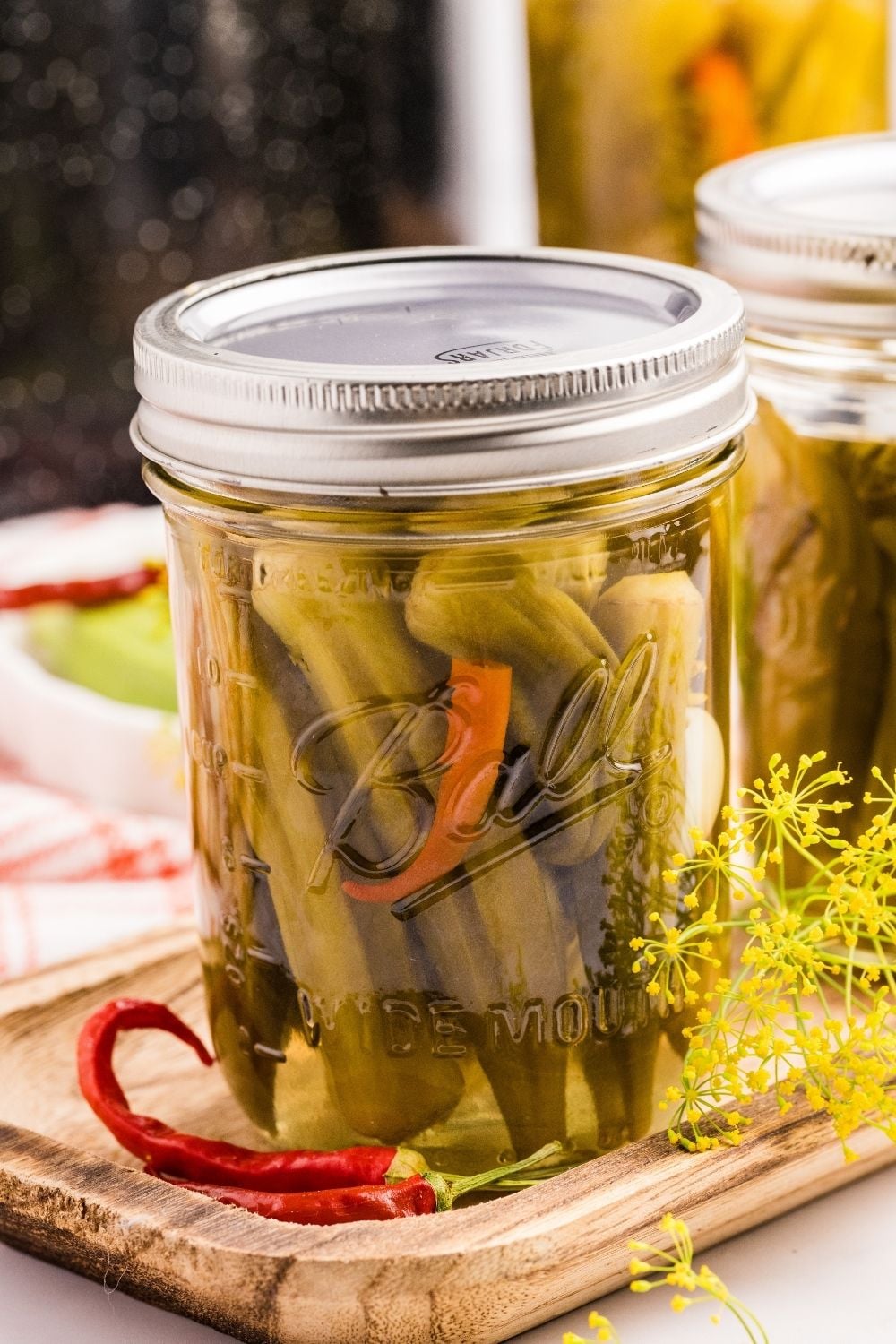 Ball mason jar of pickled okra on a wooden board with red chili peppers and fresh dill flowers.