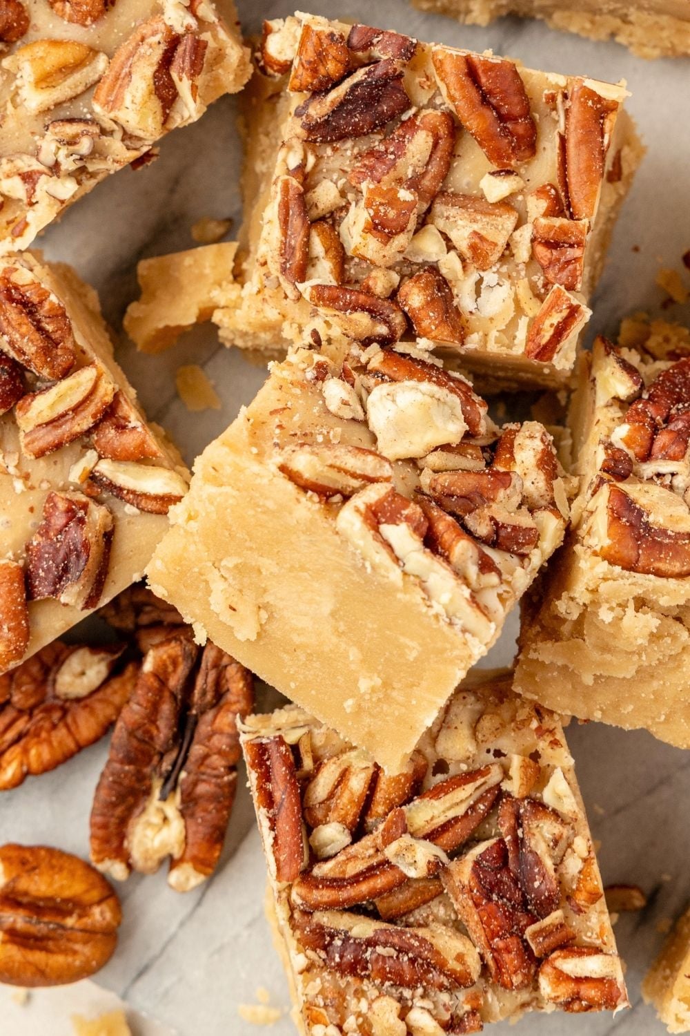 Squares of caramel-colored penuche fudge topped with chopped pecans stacked on parchment paper.