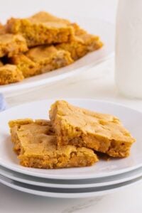 Two golden butterscotch bars stacked on white plates with more bars and a milk bottle behind.