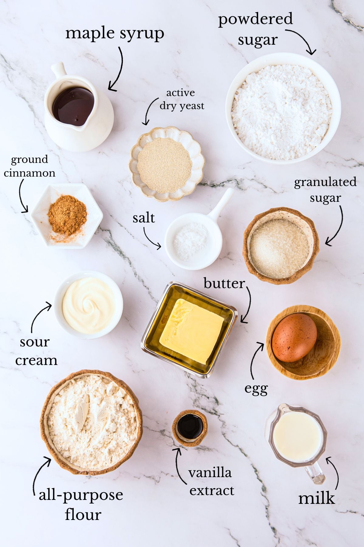 Labeled ingredients for maple bar donuts including flour, butter, egg, sour cream, yeast, cinnamon, sugar, and maple syrup on marble.