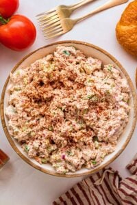 Creamy smoked chicken salad topped with paprika in a wooden bowl beside tomatoes, croissant, and gold forks.