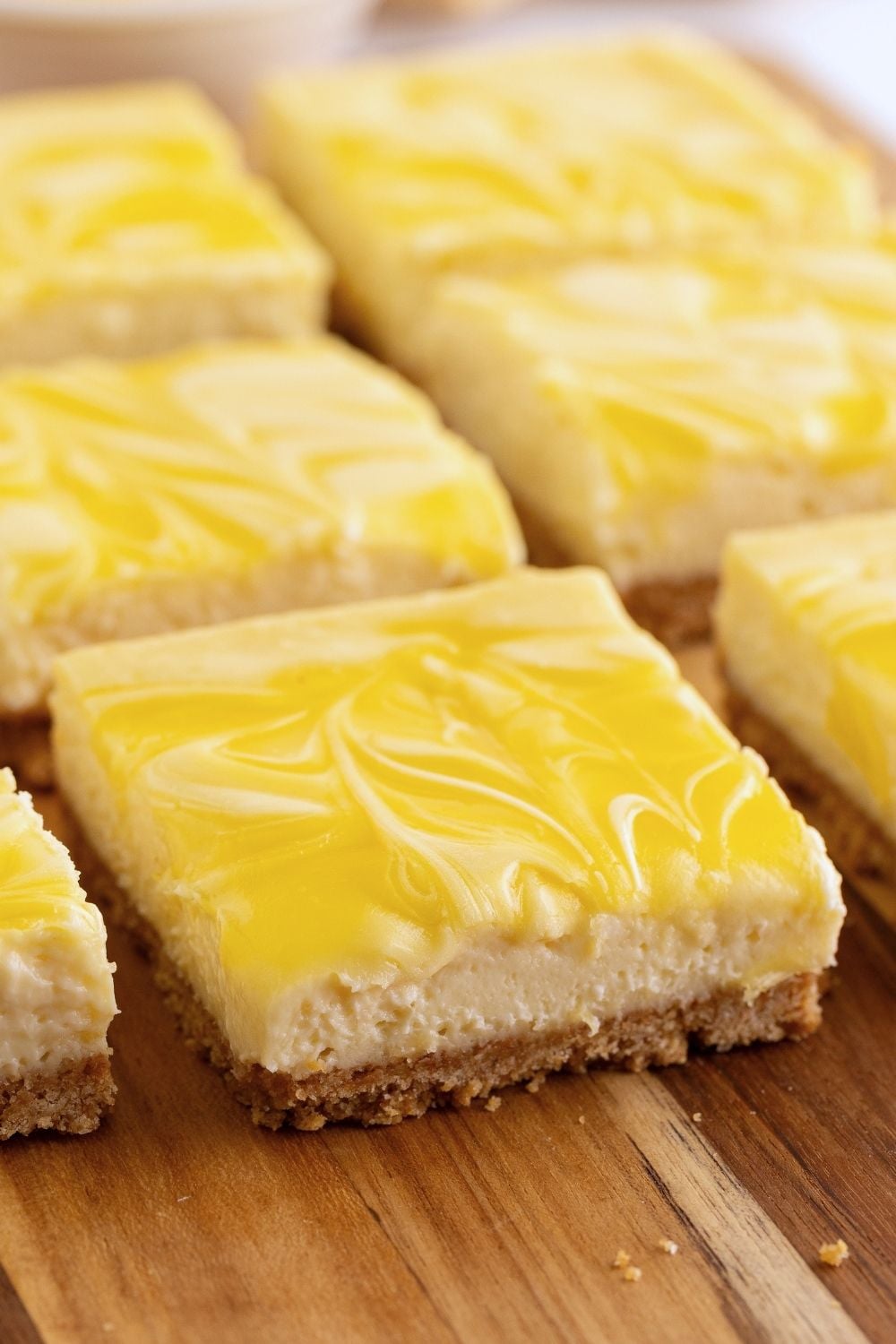 Lemon cheesecake bars with swirled yellow curd topping and graham cracker crust arranged on a wooden board.