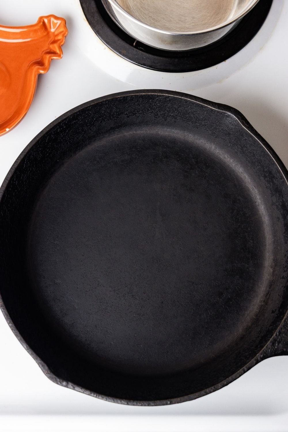 Empty seasoned cast iron skillet on a white stovetop beside an orange chicken-shaped spoon rest.
