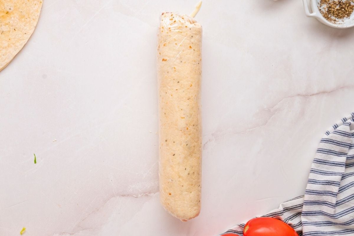 Tightly rolled tortilla wrap sealed in plastic wrap resting on marble beside striped towel and tomato.
