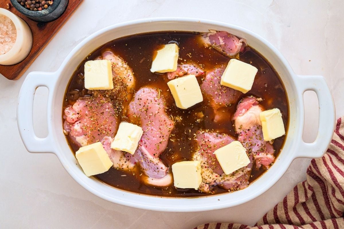 Pats of butter placed over raw seasoned chicken thighs in dark marinade inside a white baking dish.