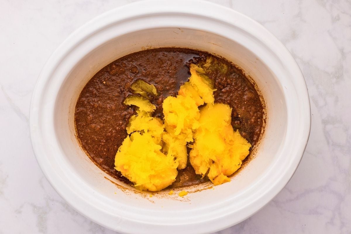 Chunks of cooked yellow pumpkin added to spiced brown sugar mixture inside a white slow cooker.
