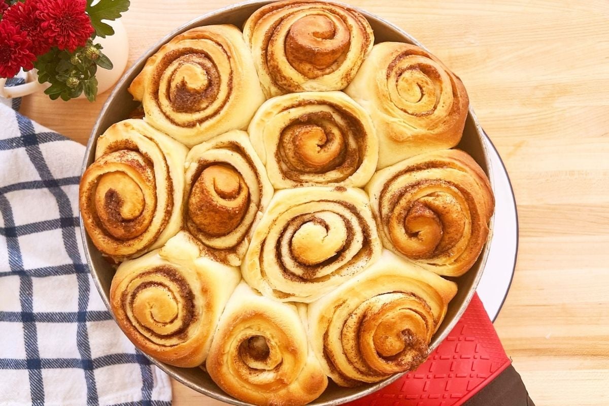 Freshly baked golden cinnamon swirl buns in a round pan beside a checkered towel and red flowers.