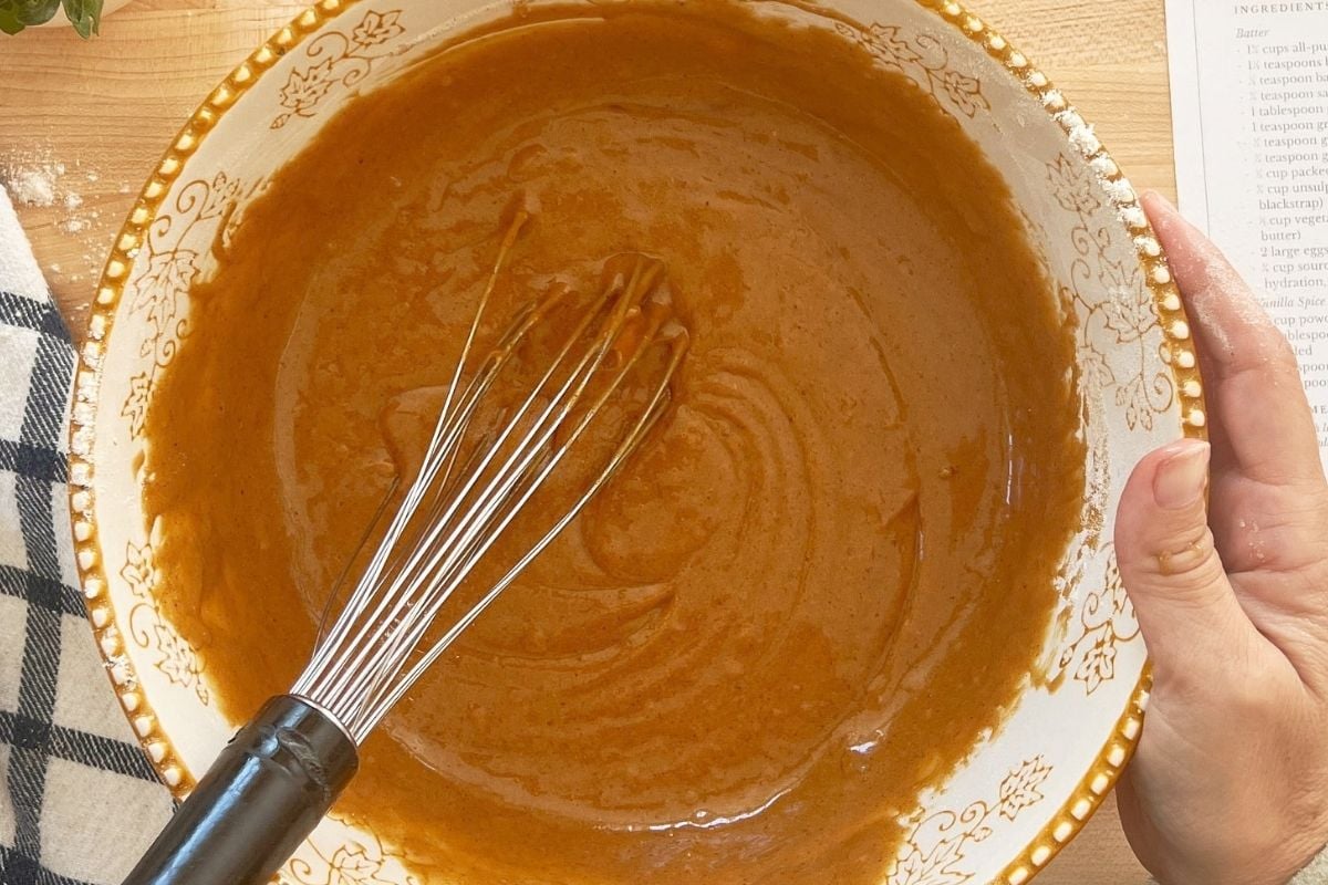Whisking smooth caramel-colored gingerbread batter with sourdough discard in a leaf-patterned ceramic bowl beside a recipe card.