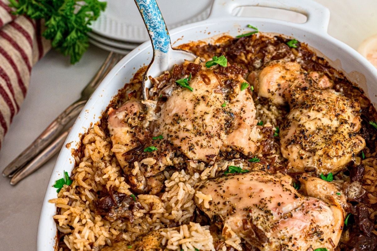 Herb-seasoned baked chicken thighs nestled in seasoned rice with parsley garnish in a white casserole dish.