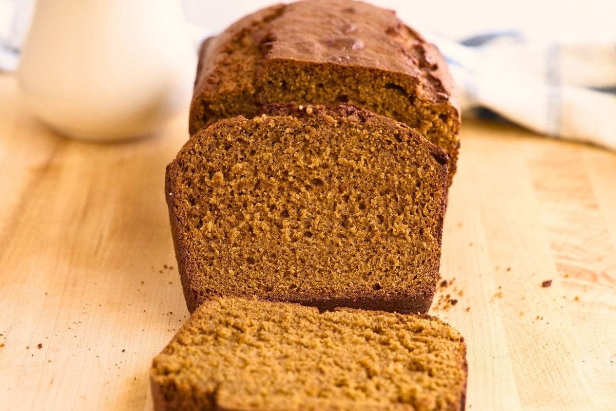 Sliced gingerbread loaf on a wooden cutting board with a milk jug and striped towel nearby.