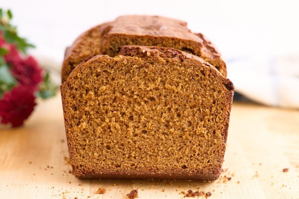 Sliced loaf of dark brown gingerbread on a wood board with red flowers blurred behind.
