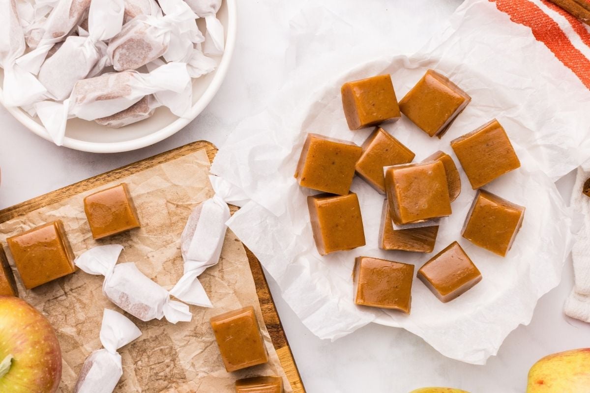 Golden-brown apple cider caramel squares on parchment-lined plate with wrapped candies and fresh apples nearby.