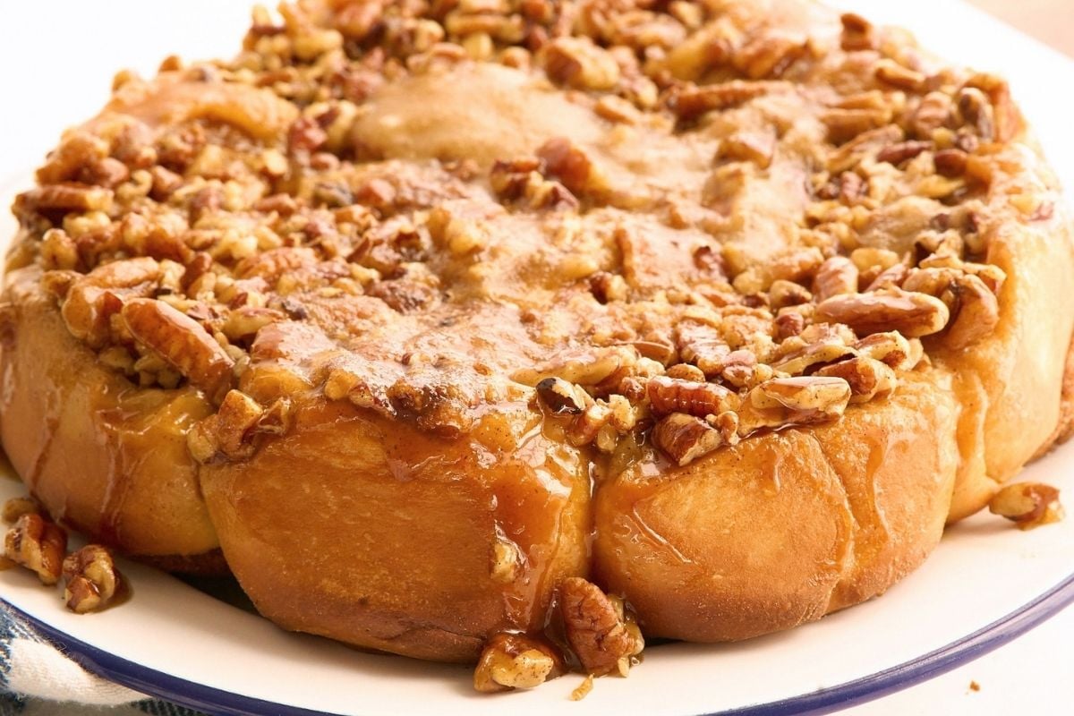 Round pan of golden sticky buns topped with caramel glaze and chopped pecans on a blue-rimmed plate.