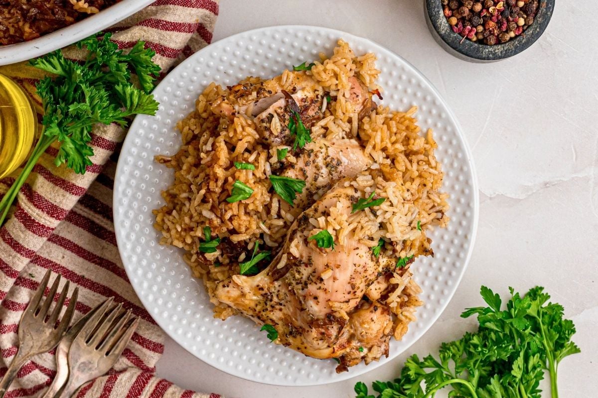 Baked chicken thighs on seasoned rice garnished with parsley on a white plate beside striped napkin.