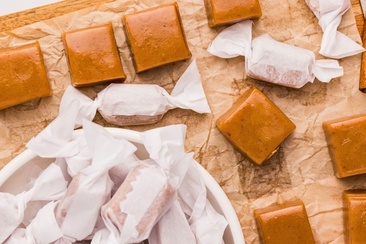 Amber apple cider caramel squares on parchment paper with some wrapped in white twisted candy papers in a bowl.
