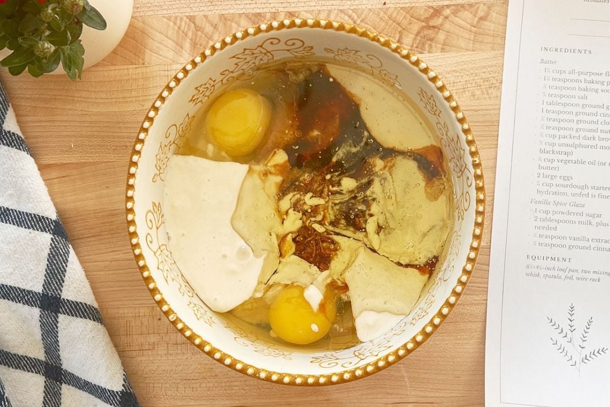 Eggs, molasses, sourdough starter, and oil pooled over flour in a gold-rimmed bowl on a wooden counter.