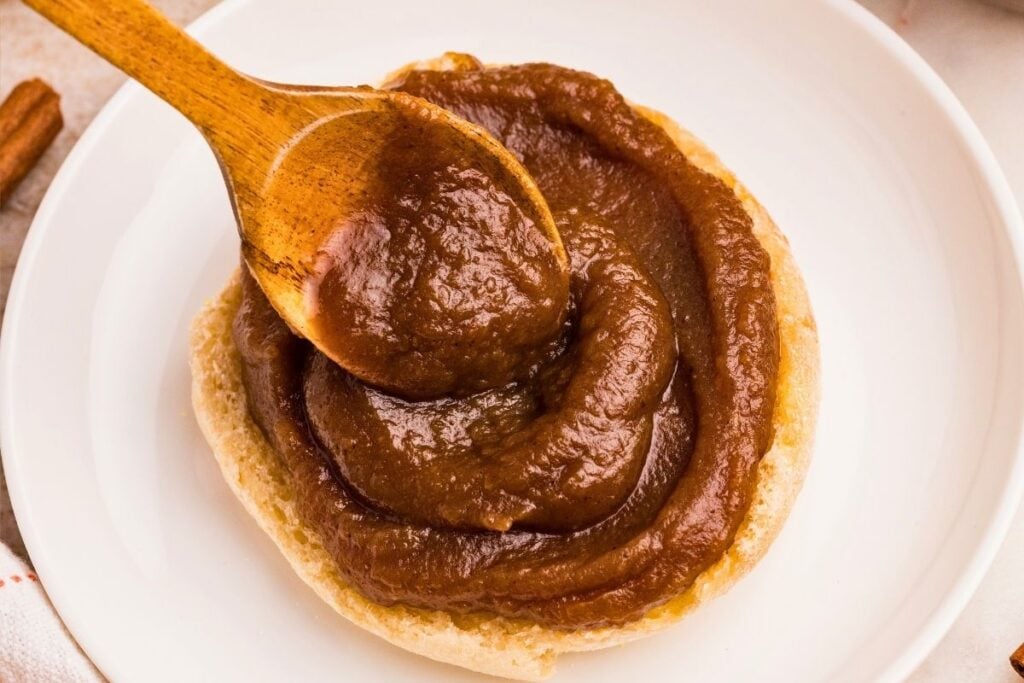 A wooden spoon spreads thick brown pumpkin butter onto a biscuit on a white plate 