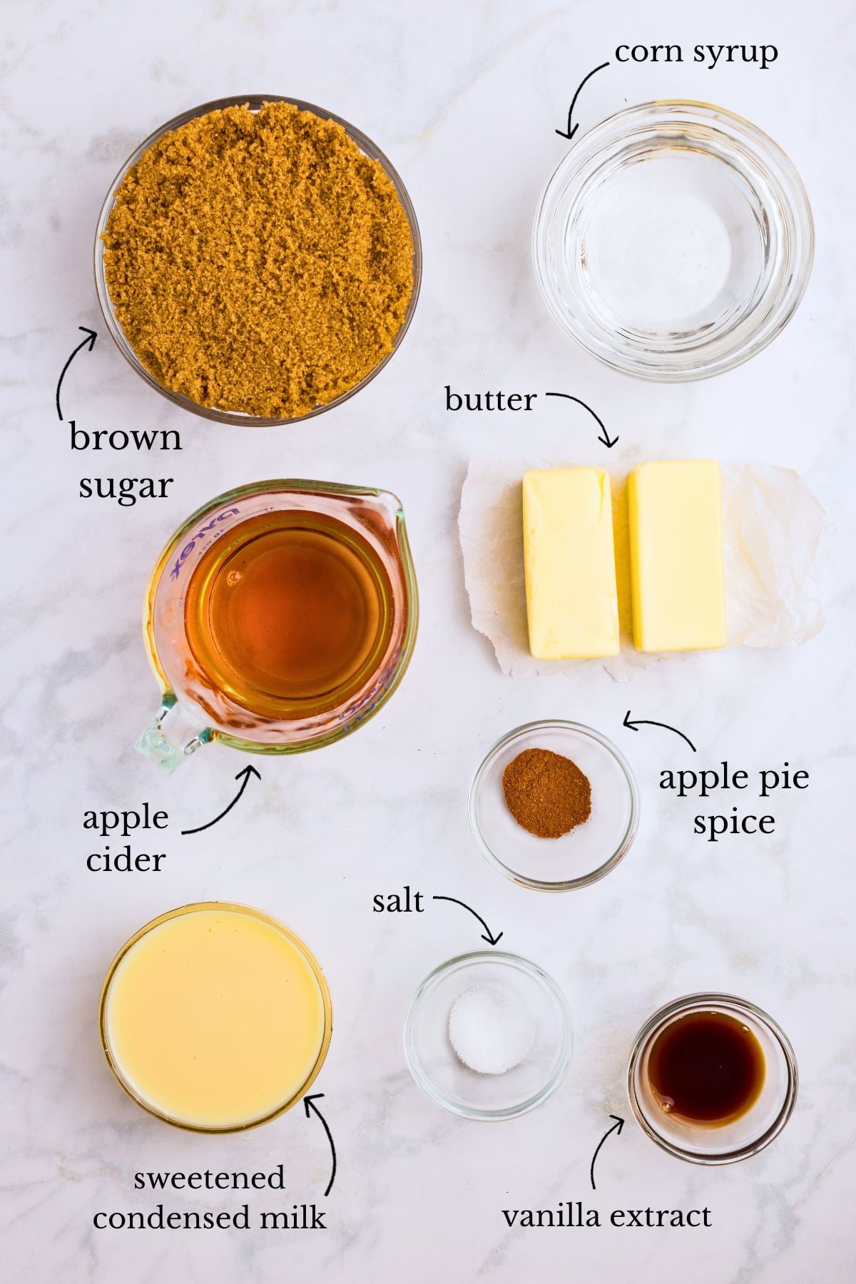 Labeled bowls of brown sugar, corn syrup, butter sticks, apple cider, apple pie spice, salt, condensed milk, and vanilla on marble.