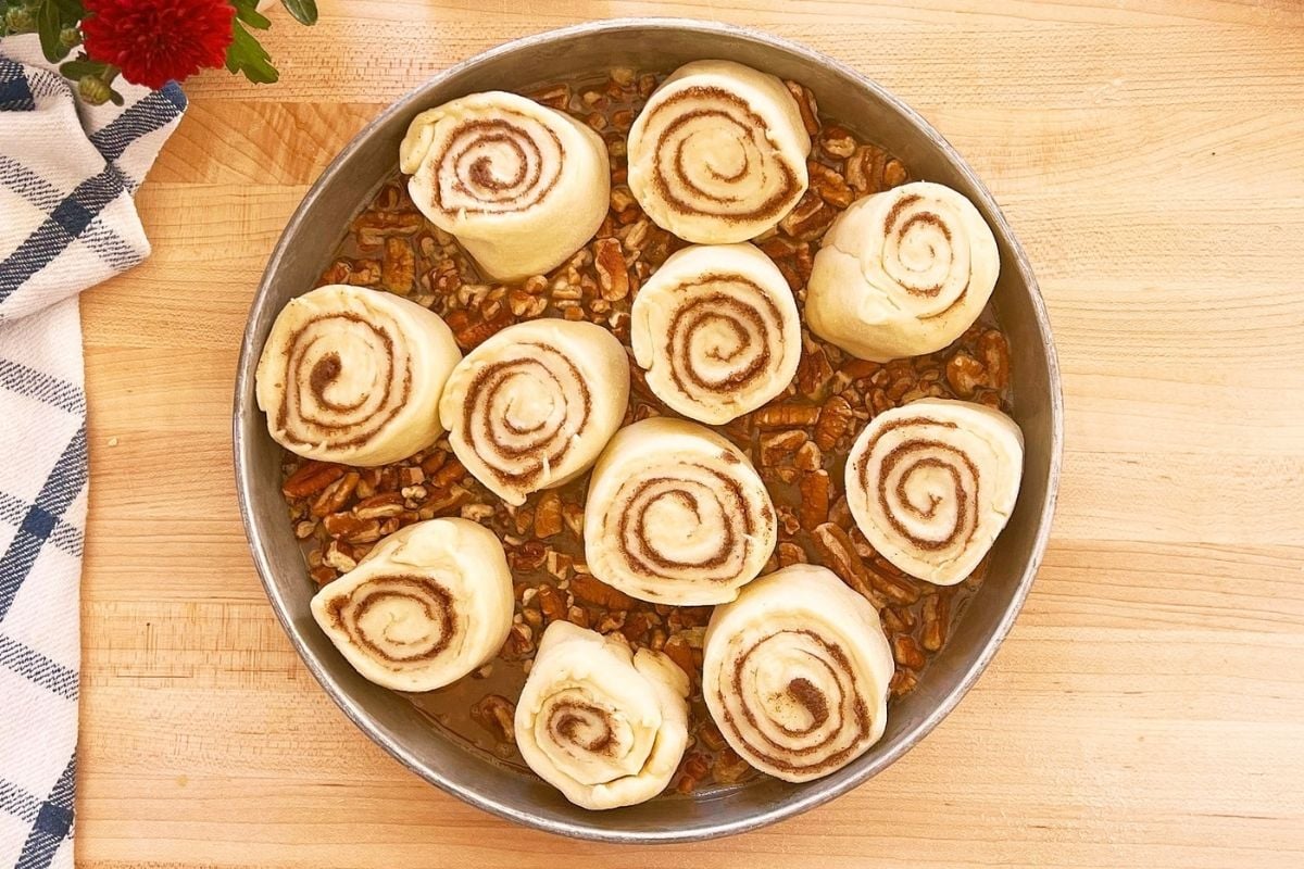 Unbaked cinnamon rolls arranged over pecans and caramel in a round metal pan on a wooden counter.