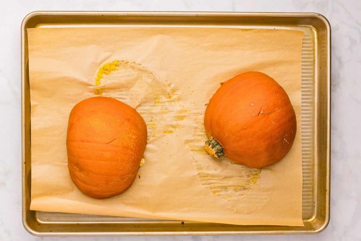 Two pumpkin halves roasting cut-side down on a parchment-lined sheet pan with released juices.