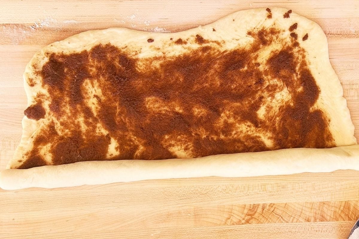 Rectangle of sourdough spread with cinnamon sugar filling being rolled up from the top edge on a wooden board.