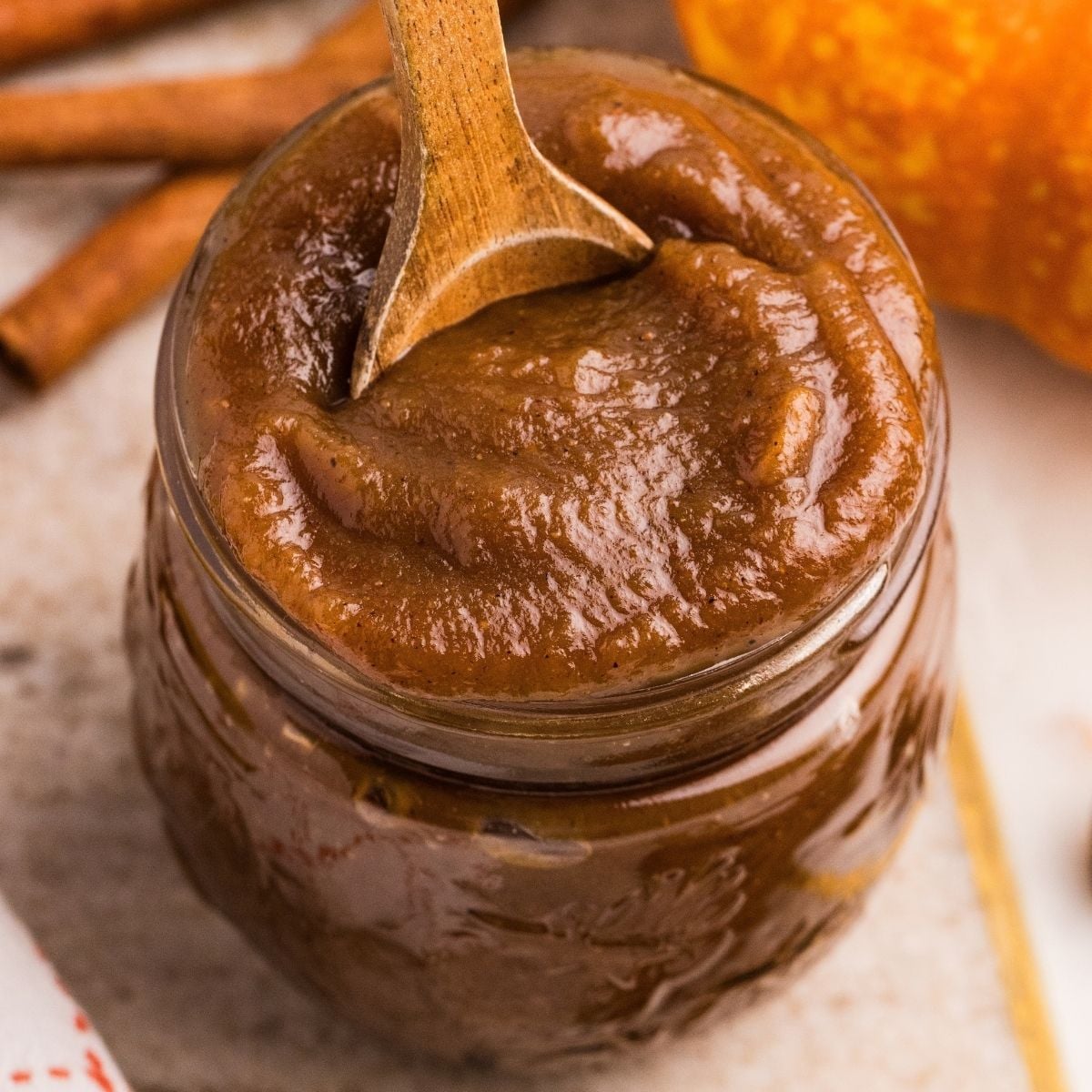 Glass mason jar filled with thick brown pumpkin butter, wooden spoon dipped in, cinnamon sticks and pumpkin nearby.