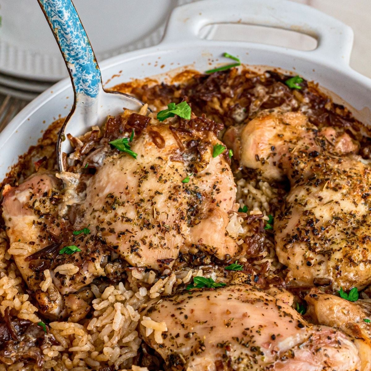 Herb-seasoned baked chicken thighs nestled over rice in a white casserole dish with blue-handled serving spoon.
