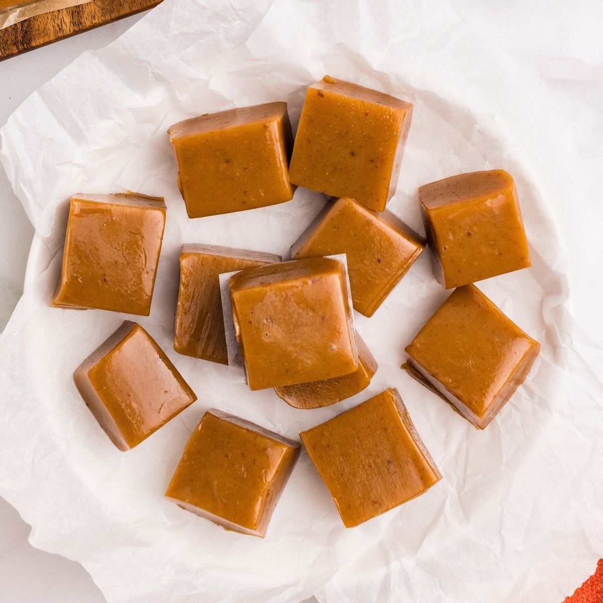Golden brown apple cider caramel squares piled on crinkled white parchment paper beside a wooden board.